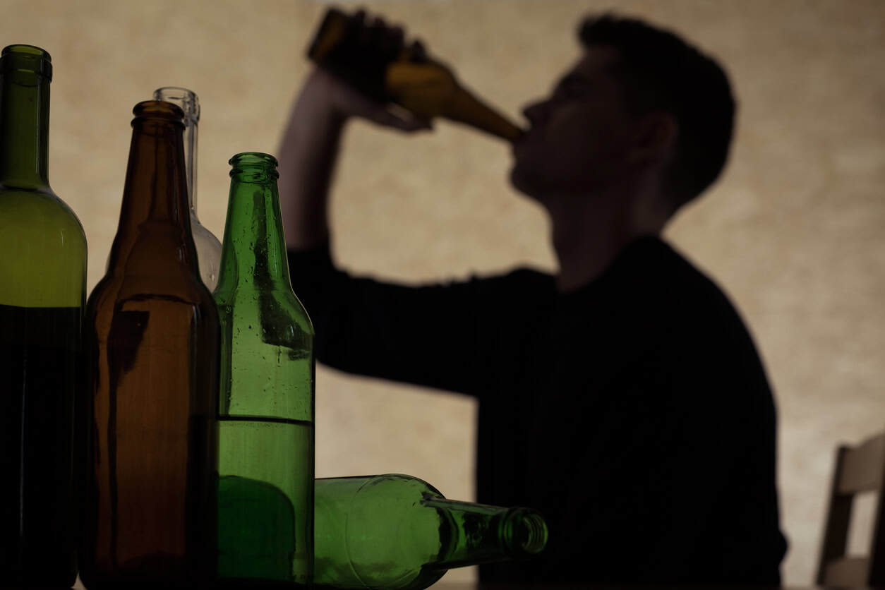 A silhouette of a person drinking from a bottle, with various empty glass bottles in the foreground