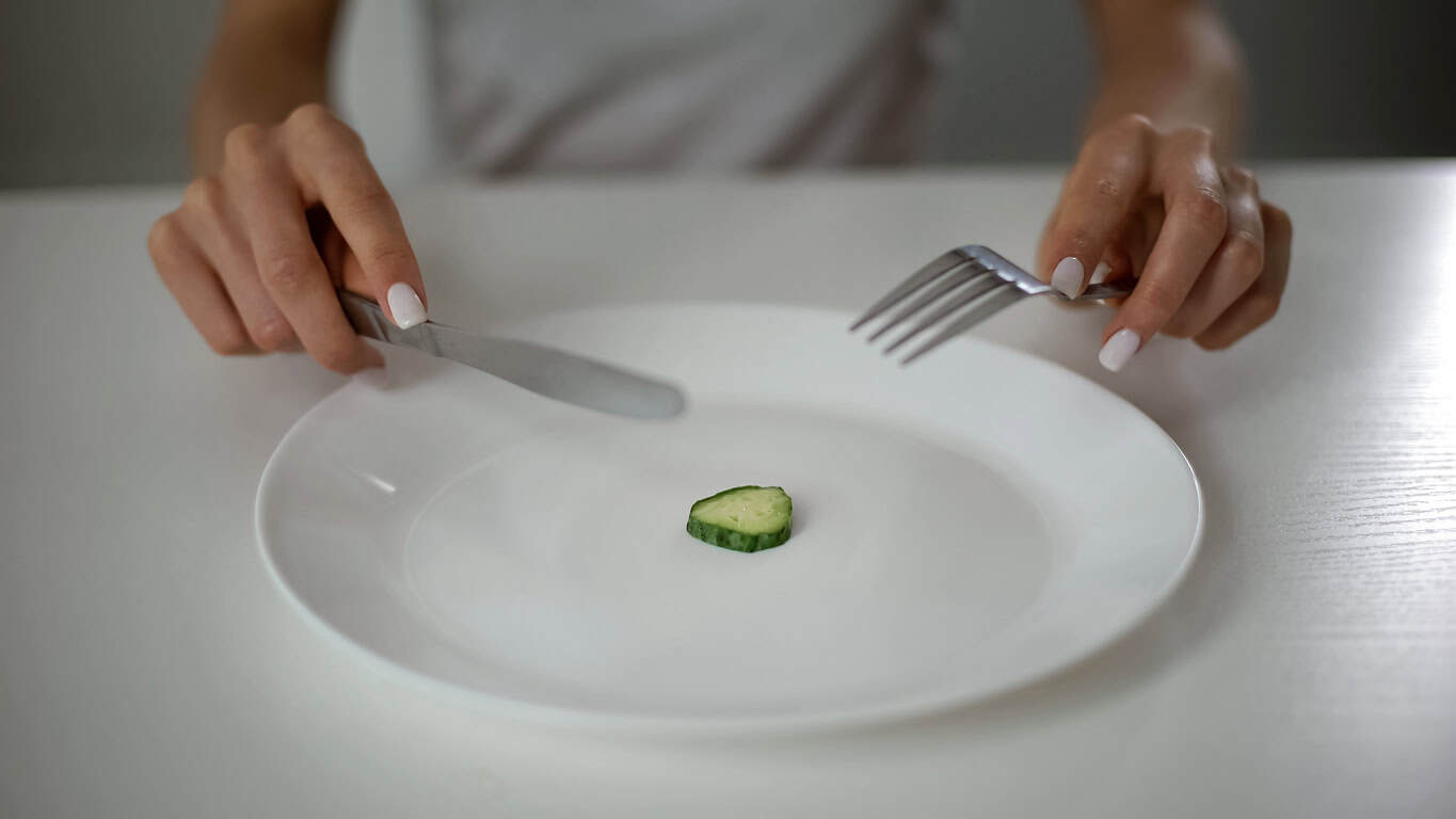 A person's hands holding a knife and fork, poised over a single slice of cucumber on a white plate