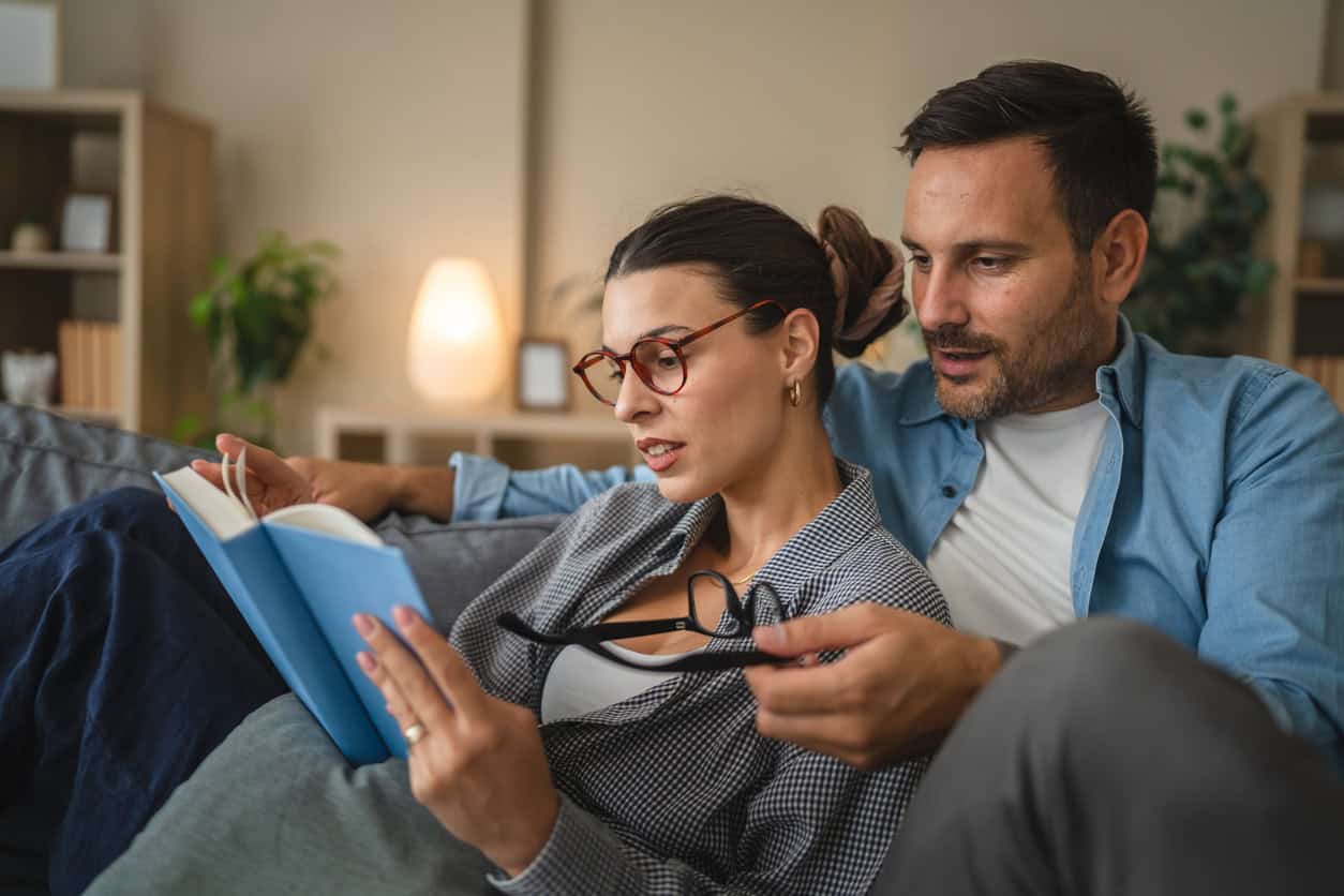 A couple sits on a couch reading a book together, with one partner holding glasses while discussing the pages